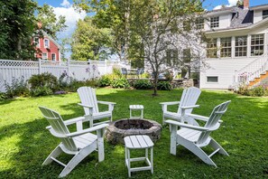 A backyard with a fire pit, four Adirondack chairs, and a white picket fence.