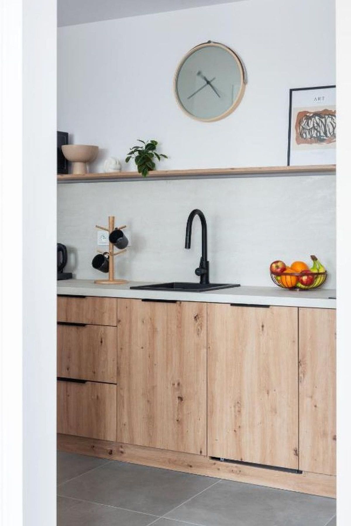 A kitchen with a black faucet, wooden cabinets, a white countertop, a clock, a bowl of fruit, and a framed picture.