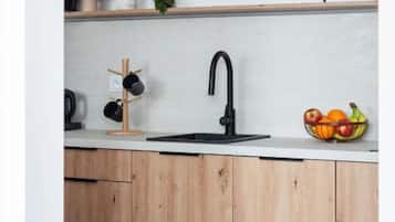 A kitchen with a black faucet, wooden cabinets, a white countertop, a clock, a bowl of fruit, and a framed picture.
