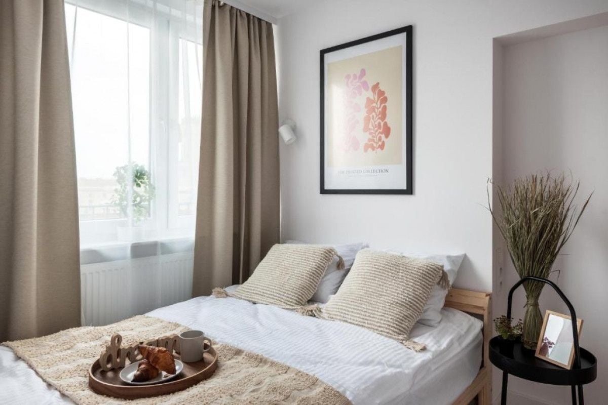 A neatly made bed with a white comforter and two pillows, a bedside table with a vase and a picture frame, and a framed artwork on the wall.