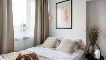 A neatly made bed with a white comforter and two pillows, a bedside table with a vase and a picture frame, and a framed artwork on the wall.