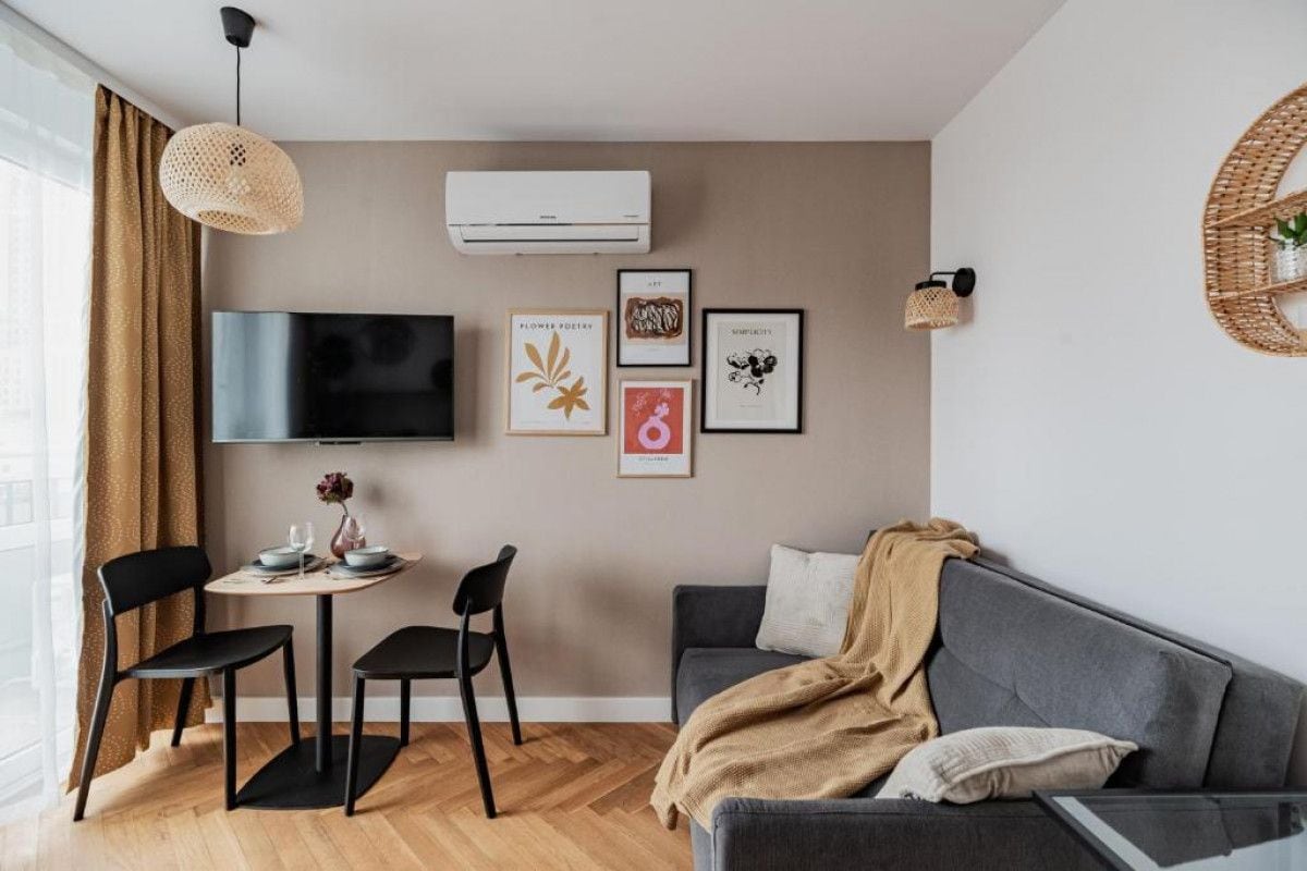 Ein modernes Wohnzimmer mit einem grauen Sofa, einem Couchtisch und einem an der Wand befestigten Fernseher. An der Wand befinden sich gerahmte Kunstwerke und eine Pendelleuchte.