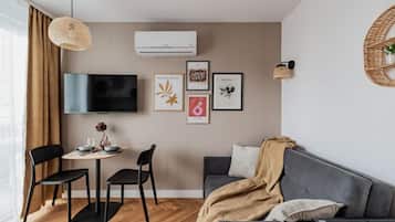 Ein modernes Wohnzimmer mit einem grauen Sofa, einem Couchtisch und einem an der Wand befestigten Fernseher. An der Wand befinden sich gerahmte Kunstwerke und eine Pendelleuchte.