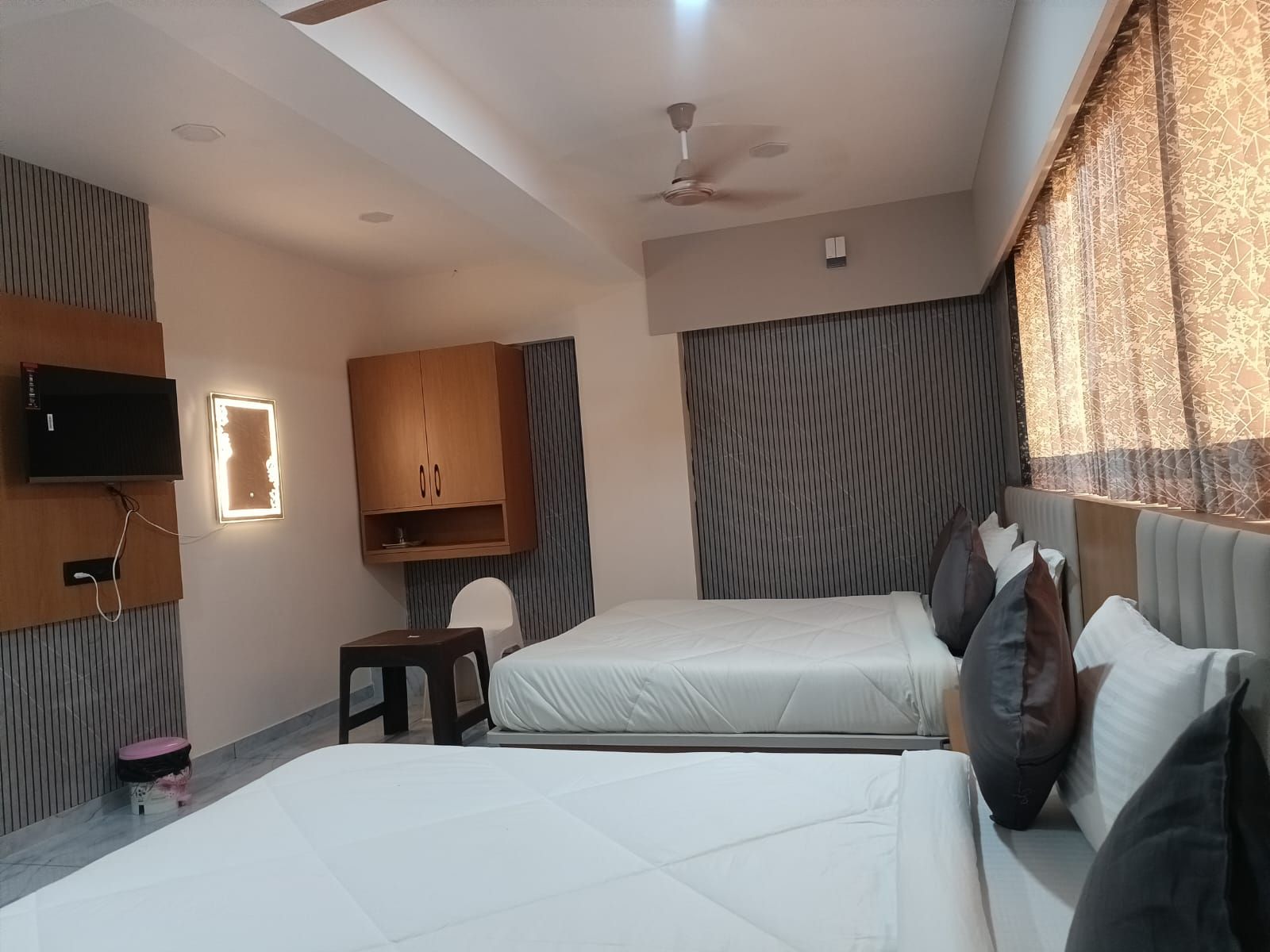 A hotel room with two beds, a TV mounted on the wall, a ceiling fan, and a window with patterned curtains.