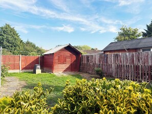 Un abri de jardin en bois peint en rouge, situé dans une cour arrière délimitée par une clôture en bois, et entouré d’herbe verte et de plantes jaunes.