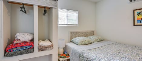 A bedroom with a bed, a nightstand, a lamp, a window with blinds, a wardrobe, and a wall-mounted air conditioner.