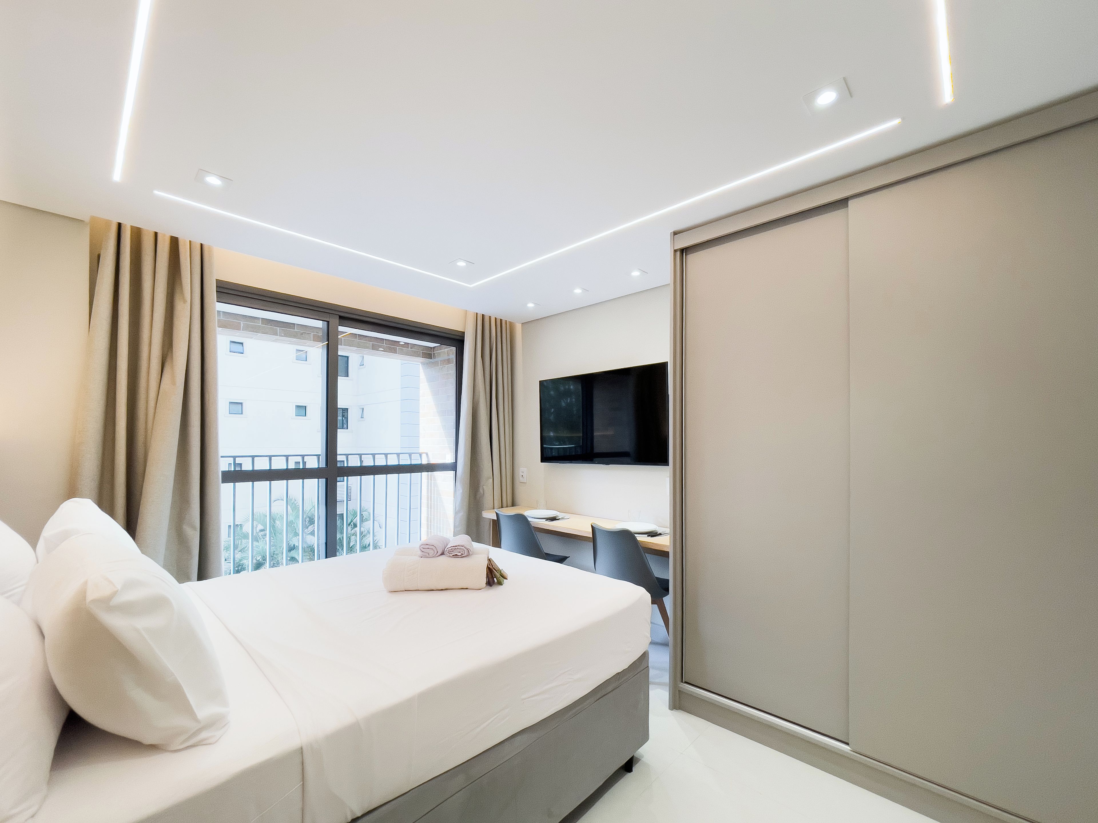 A modern hotel room with a bed, a desk, a chair, a TV, and a view of the outside.