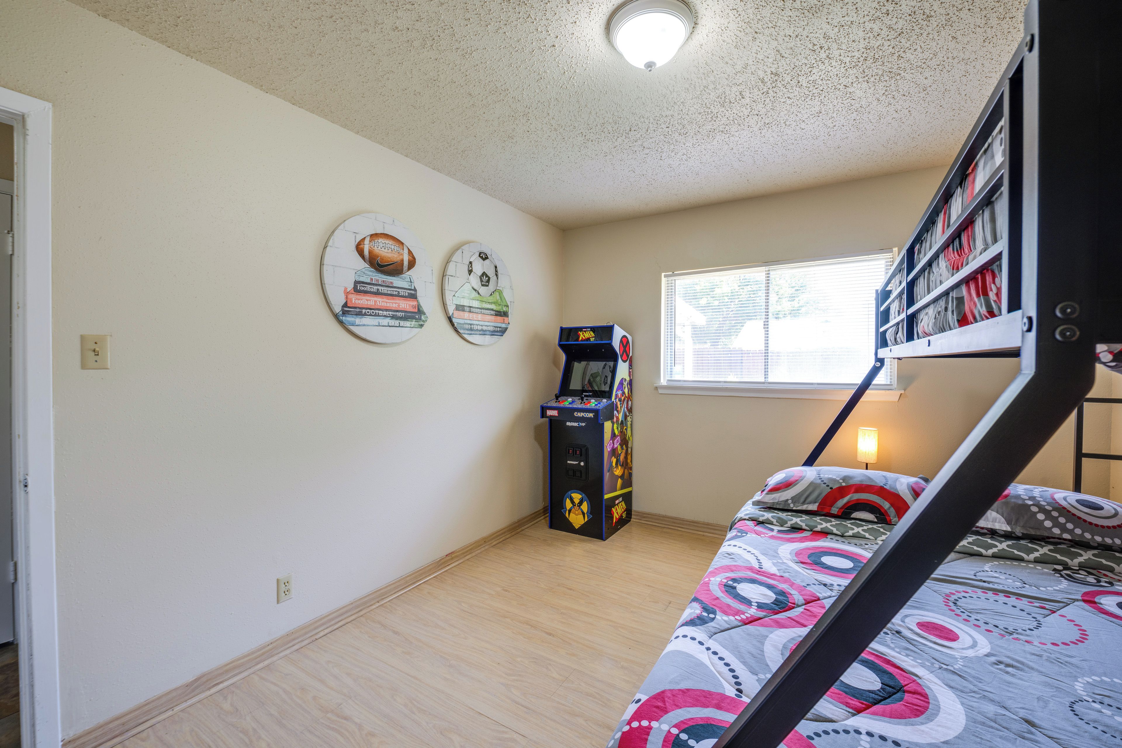 Une chambre avec un lit superposé, un ventilateur de plafond, une fenêtre avec des stores et une borne d’arcade fixée au mur.