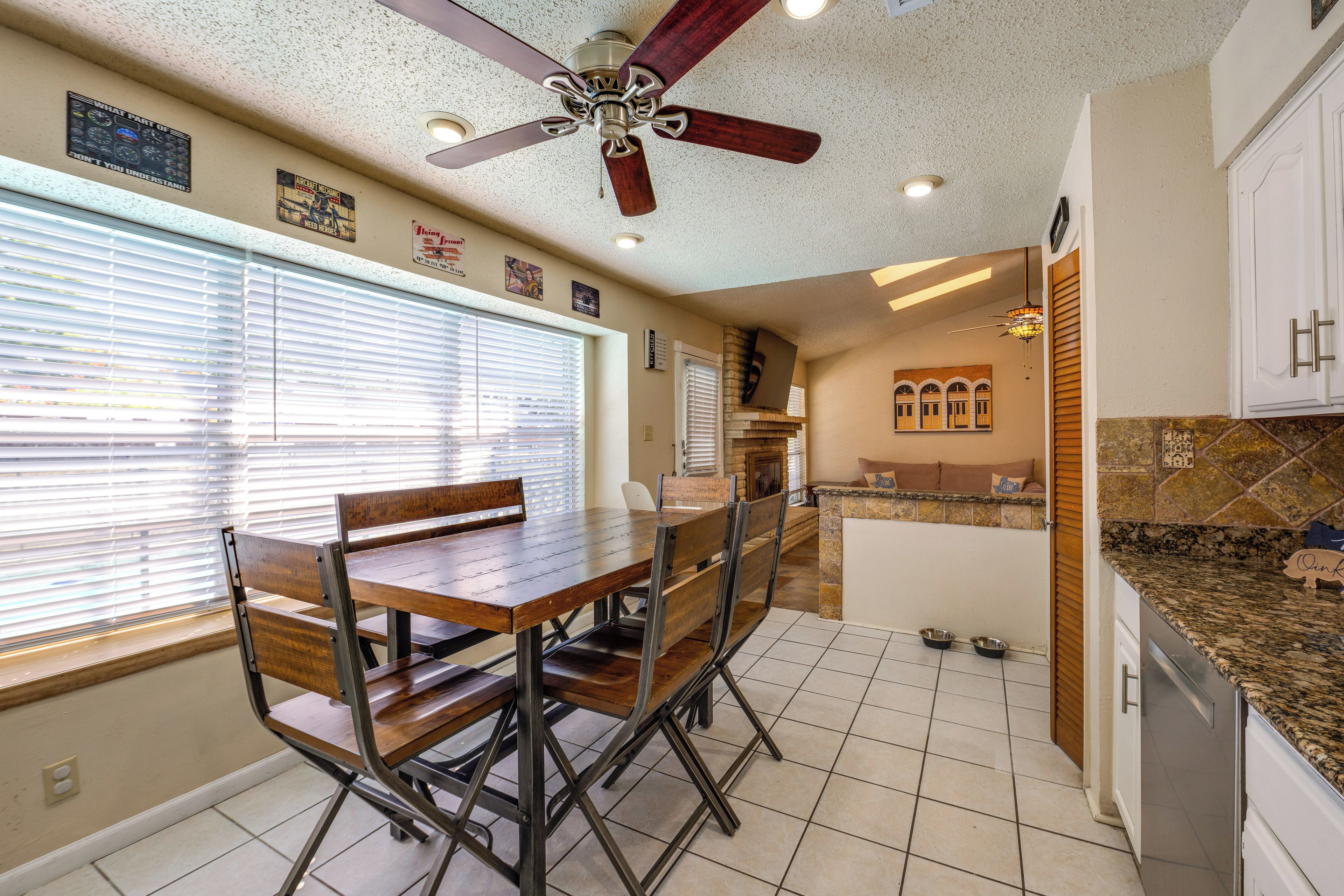 A dining area with a wooden table and chairs, a ceiling fan, and a kitchen with a stone backsplash.