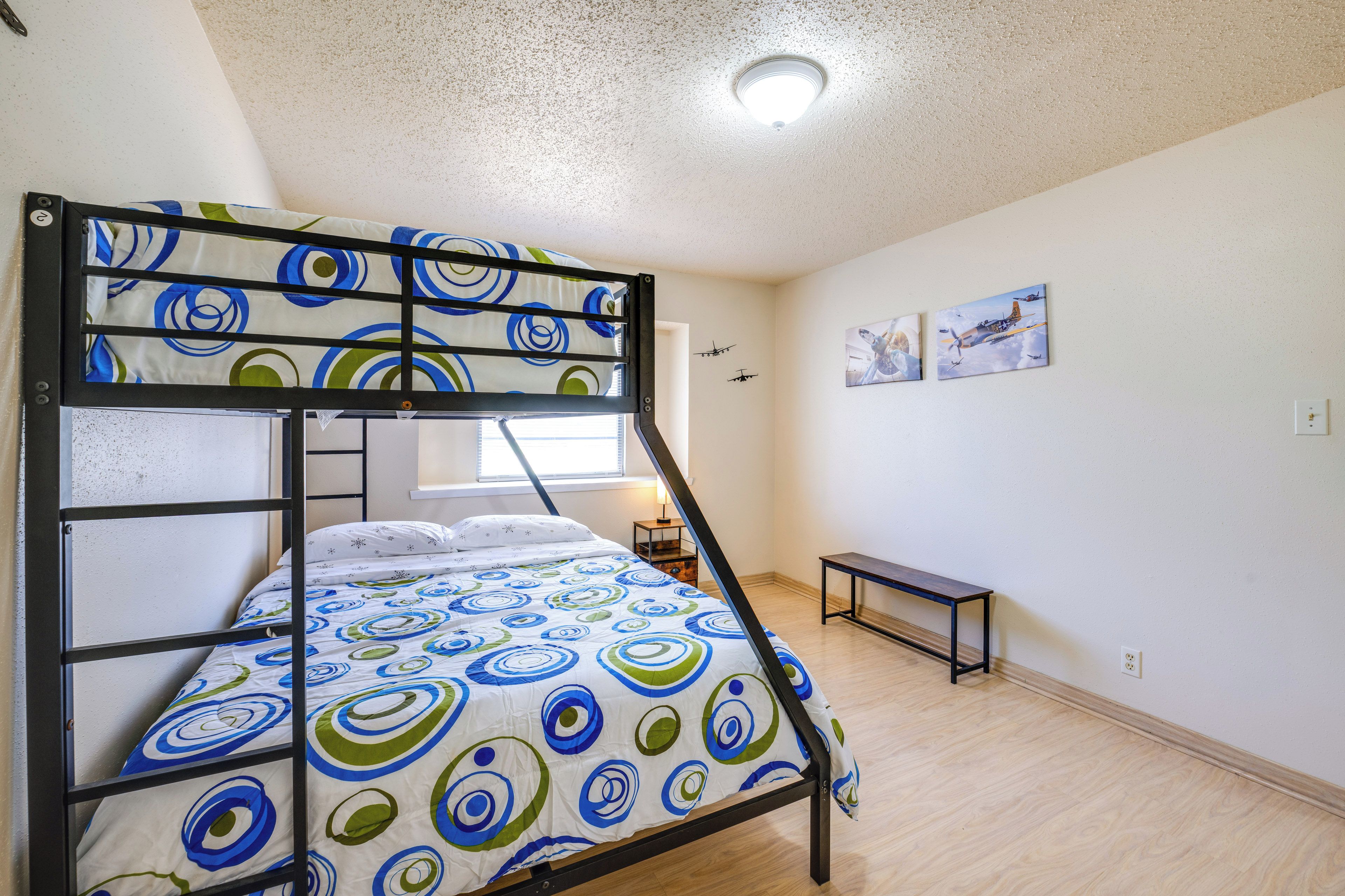 Una cama litera con una colcha estampada, una mesita de noche pequeña, un banco y dos cuadros en la pared.