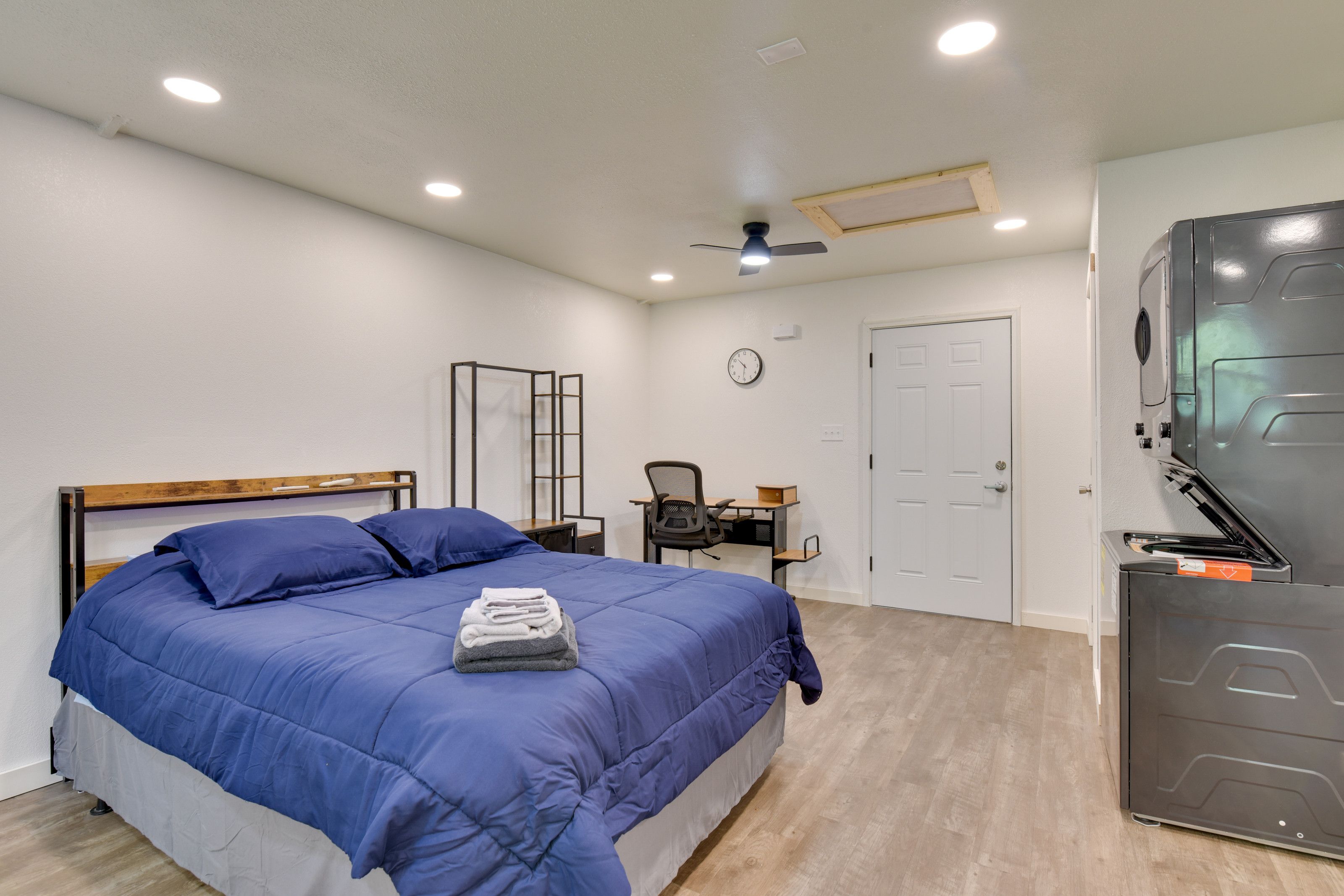A bedroom with a bed, a desk, a chair, a ceiling fan, a clock, and a door.