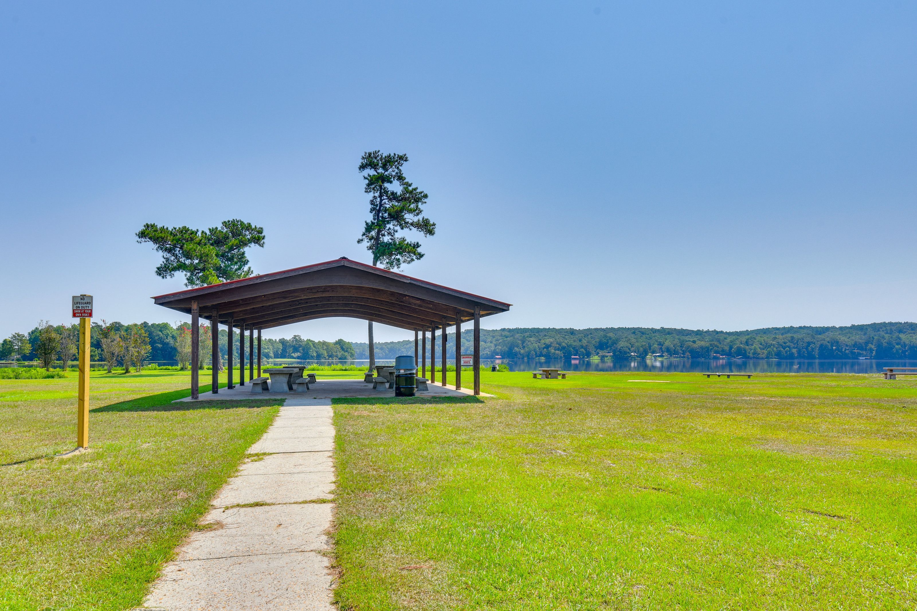 A covered picnic area with benches and a grill, surrounded by a grassy field and trees, with a lake in the background.