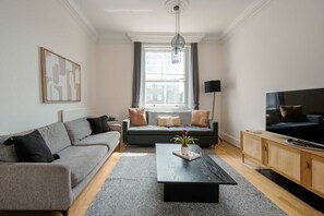A modern living room with a grey sofa, a coffee table with a plant, a TV on a wooden cabinet, and a large window with curtains. - South Kensington 2-bed - Lift - Sleeps 6 (London)