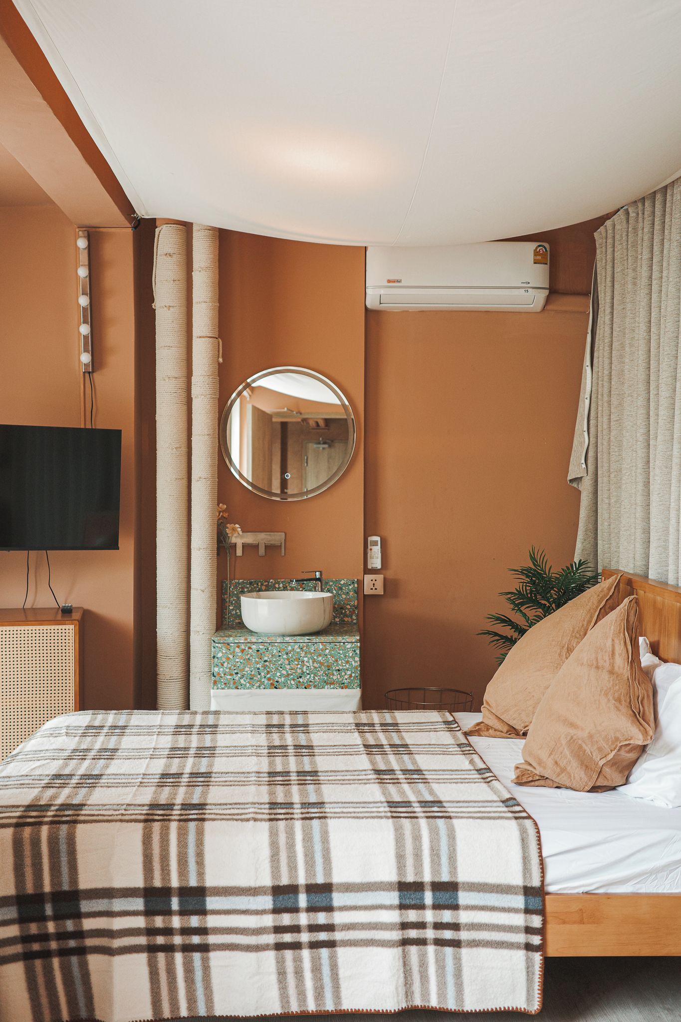 A hotel room with a bed, a round mirror, a television, a sink, and an air conditioning unit.