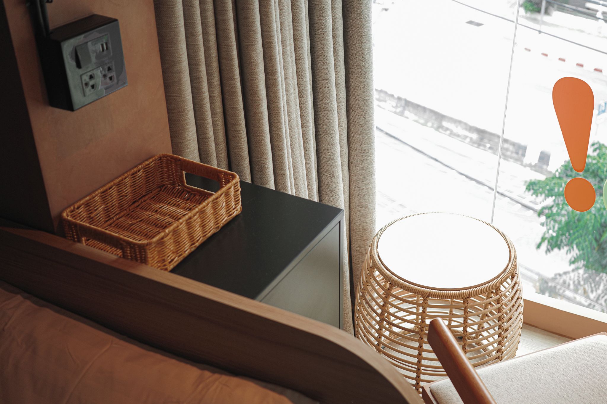 Una habitación con cama, mesita de noche, enchufe, ventana con cortinas y vista a la calle.