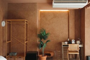 A room with a wooden wardrobe, a potted plant, a desk with a chair, a small table, a clock, and an air conditioner.