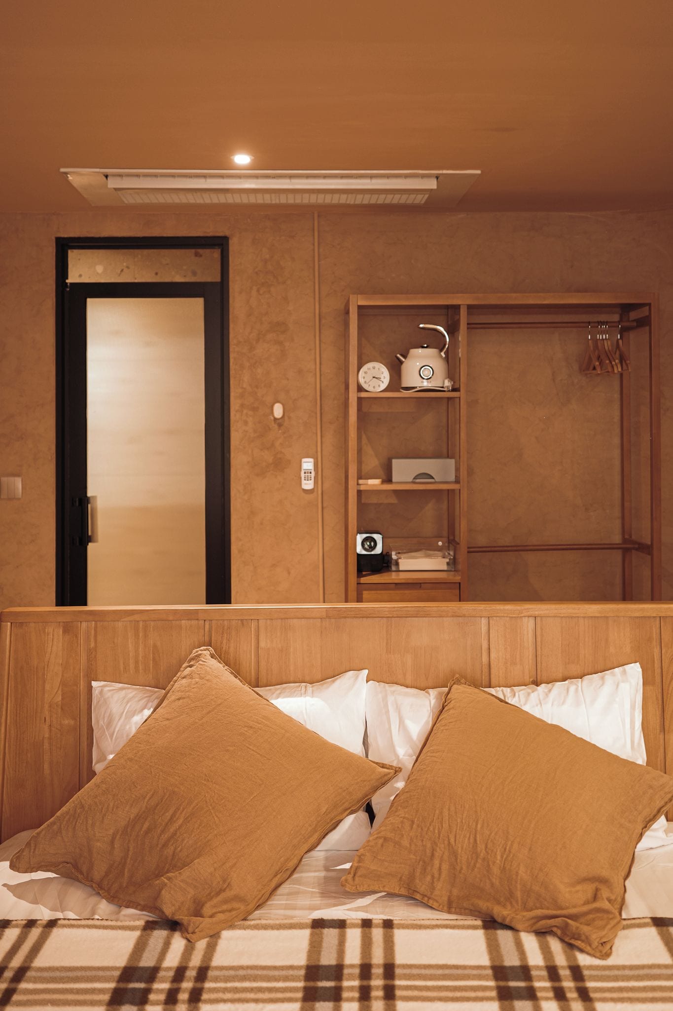A hotel room with a bed, a wooden headboard, a shelf with kitchen appliances, and a door.