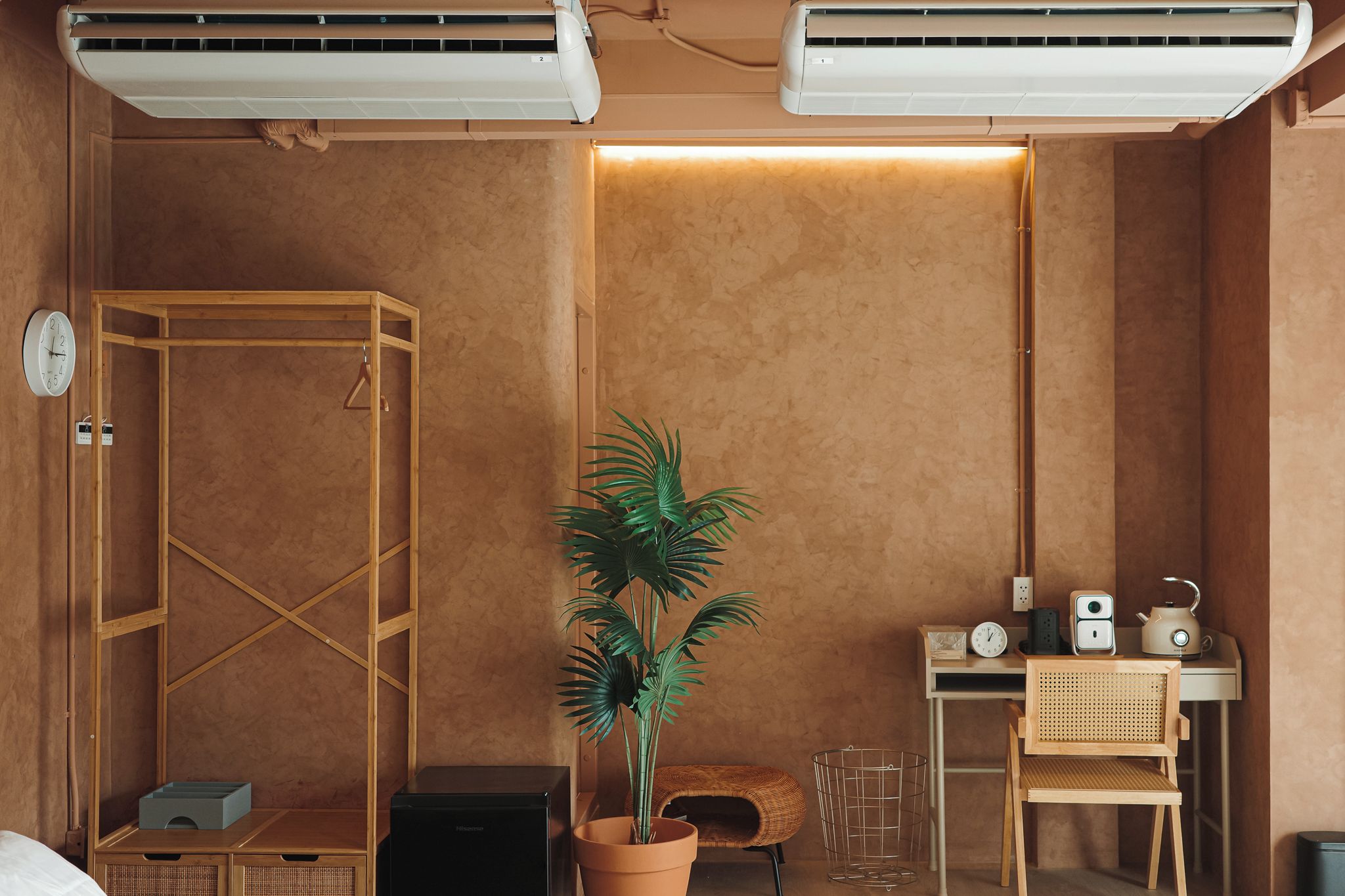 A room with a wooden wardrobe, a potted plant, a small table with a kettle, and a wall-mounted clock.
