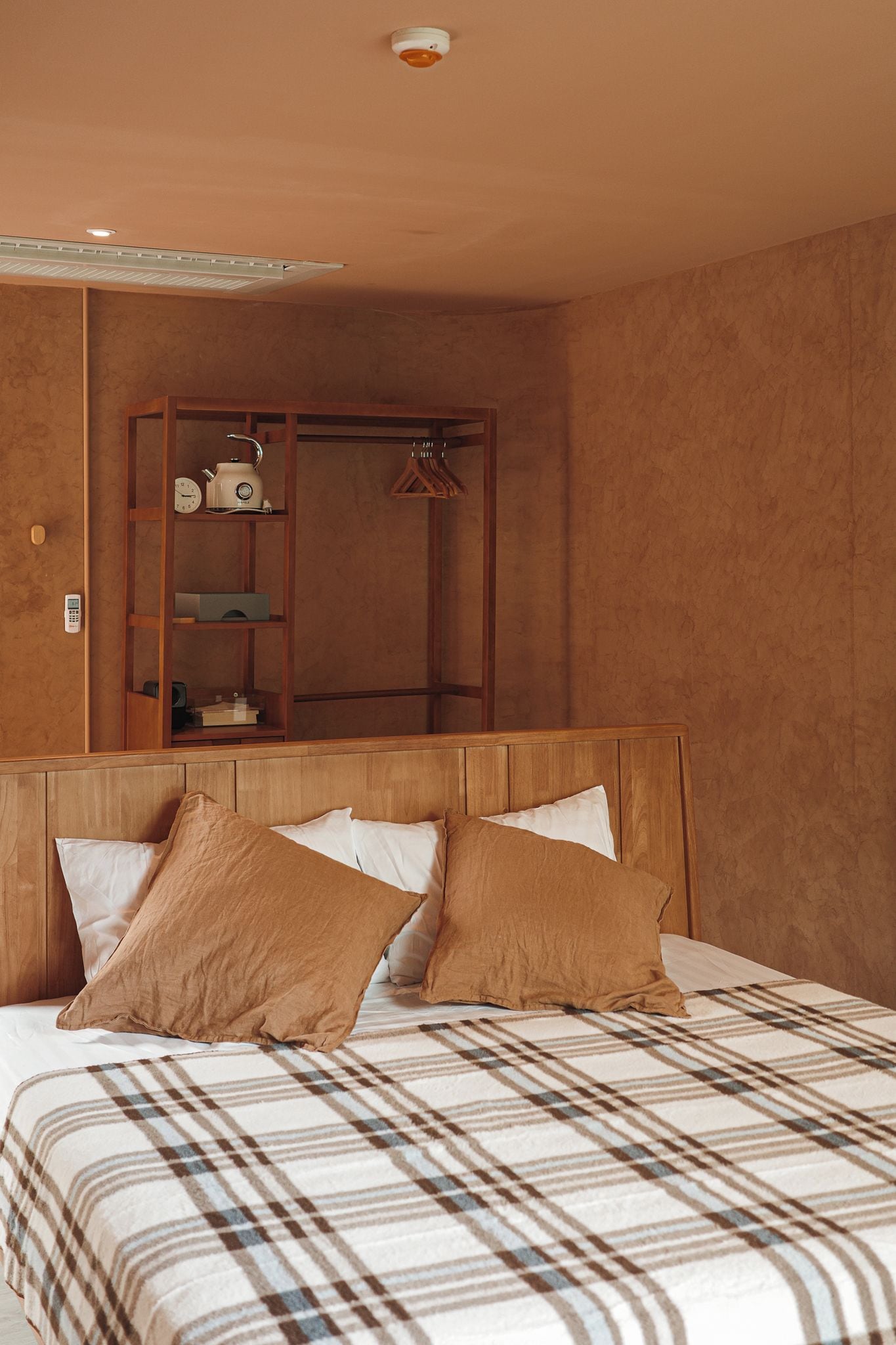 Een hotelkamer met een bed, een houten hoofdbord, een garderobe en een klein plankje met een waterkoker en een klok.