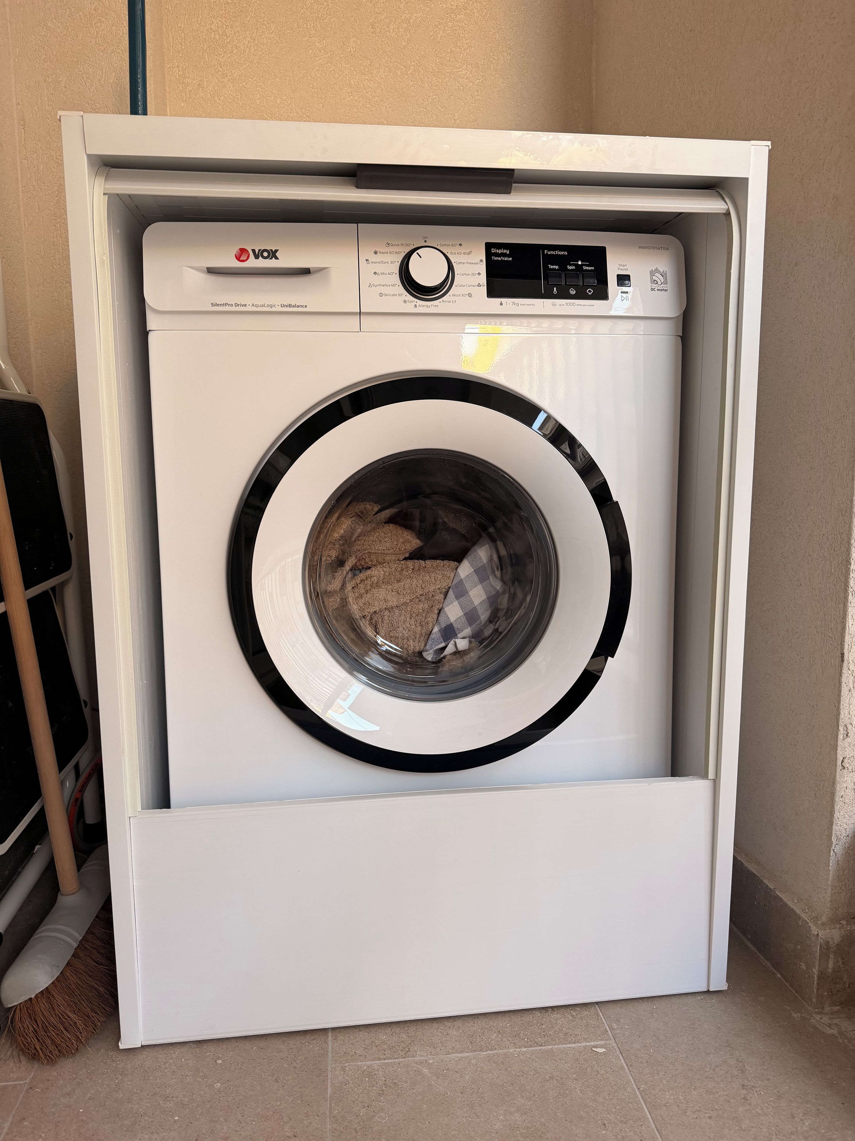 Eine kompakte, weiße Waschmaschine mit Frontlader und Bedienfeld steht in einer Raumecke mit Fliesenboden.