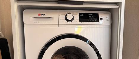 A compact, white front-loading washing machine with a control panel, placed in a corner of a room with tiled flooring.