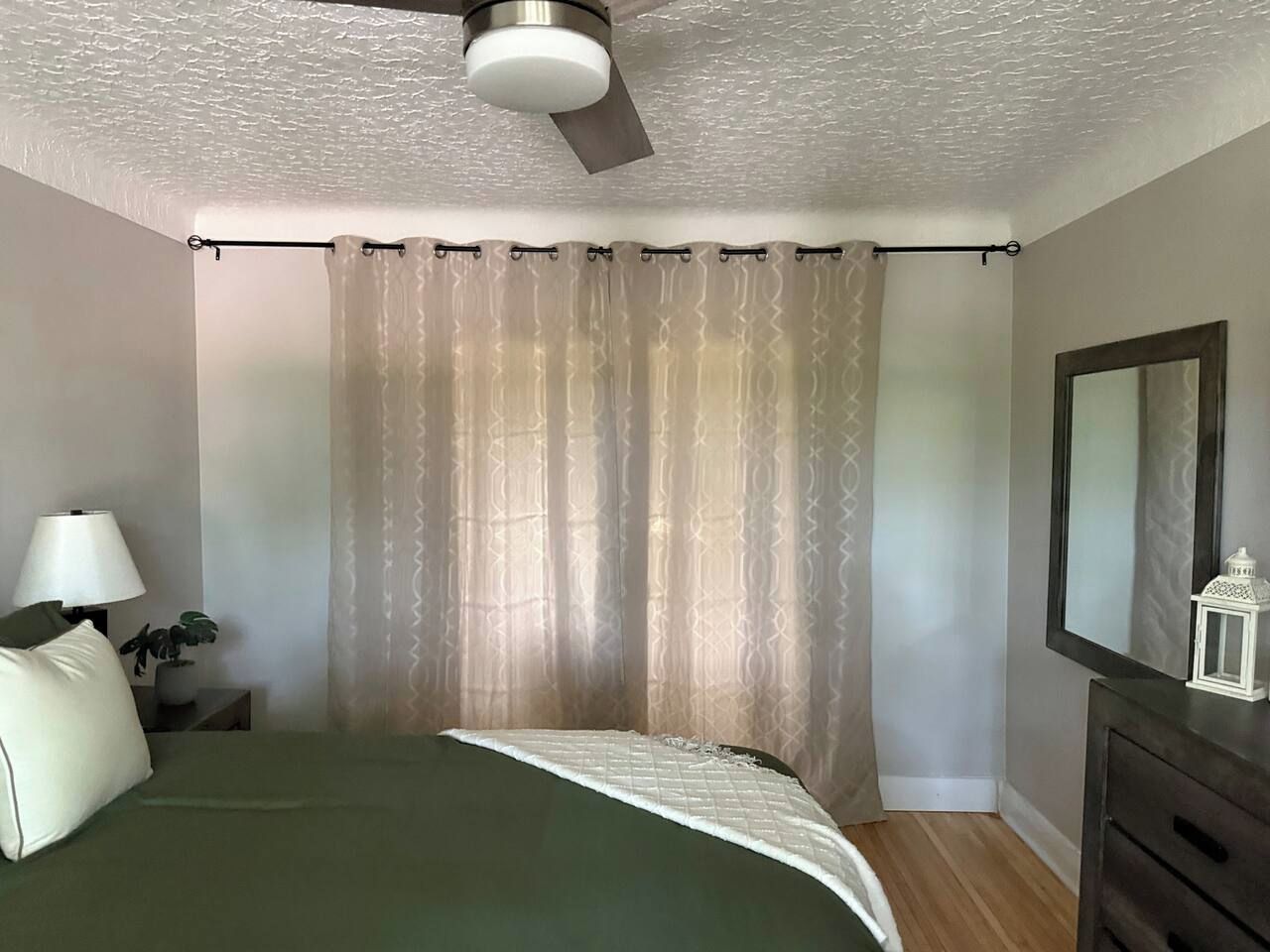 Ein Schlafzimmer mit Bett, Nachttisch, Lampe, Deckenventilator, Spiegel und Fenster mit Vorhängen.