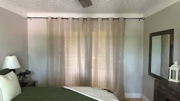 Ein Schlafzimmer mit Bett, Nachttisch, Lampe, Deckenventilator, Spiegel und Fenster mit Vorhängen.