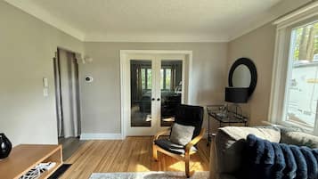 A living room with a wooden floor, a black leather sofa, a wooden chair, a glass door leading outside, and a window with a view of trees.