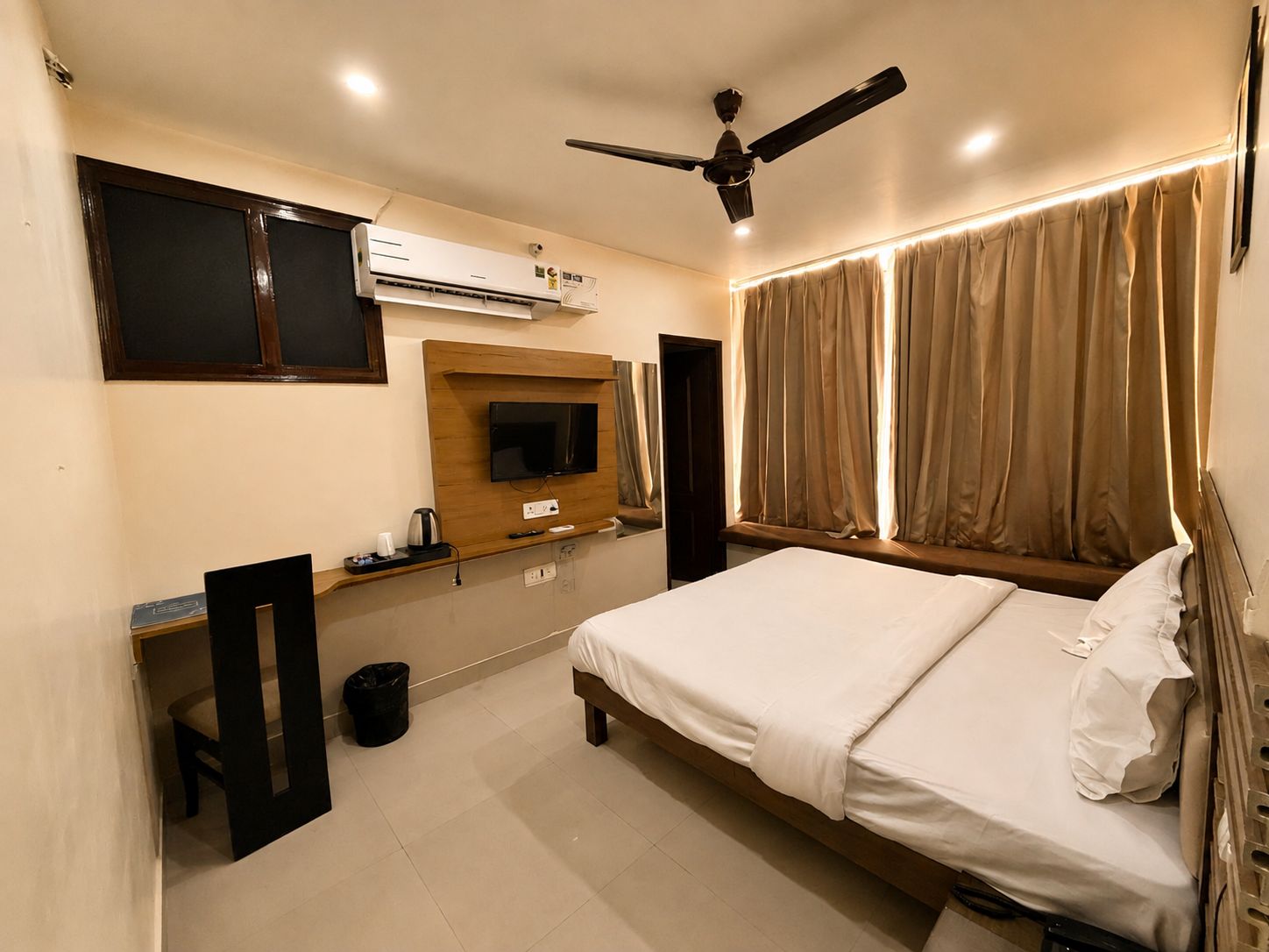 Habitación de hotel con cama, televisor, equipo de aire acondicionado, ventilador de techo y cortinas.