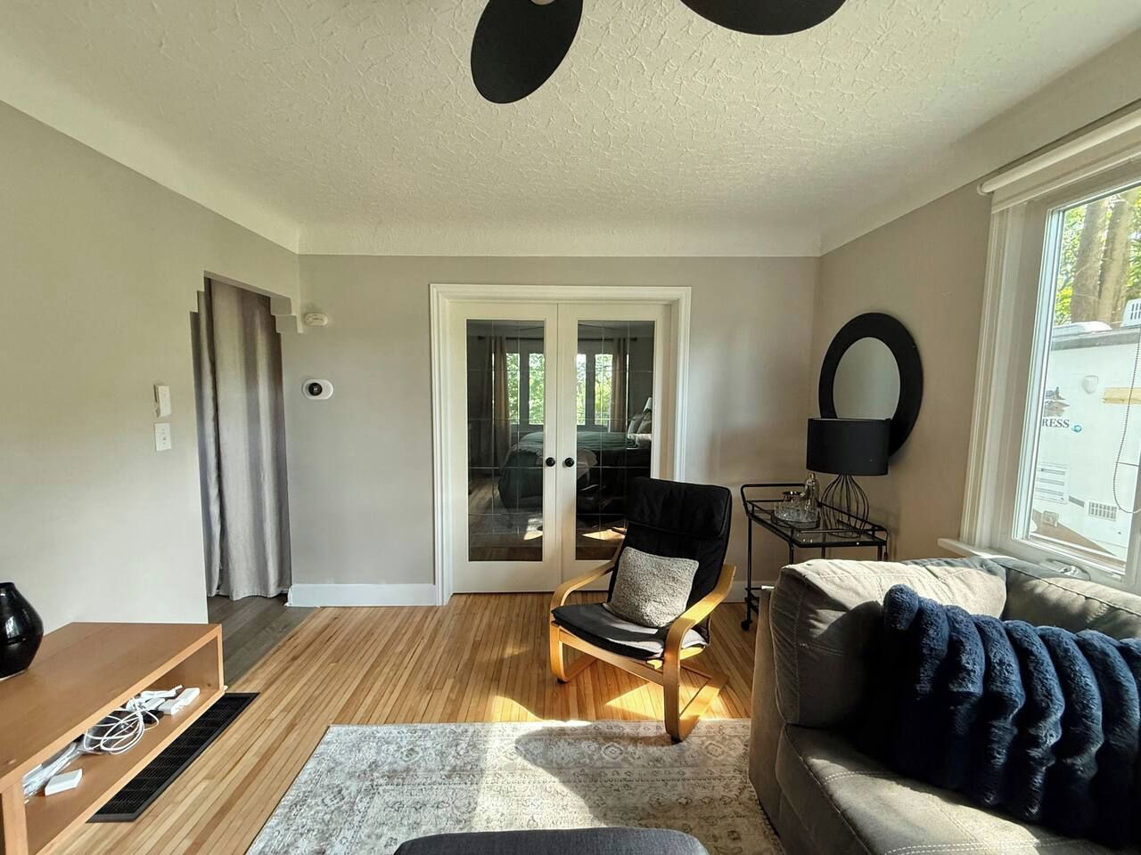 Een woonkamer met een houten vloer, een zwarte leren bank, een houten stoel, een glazen deur naar buiten en een raam met uitzicht op bomen.