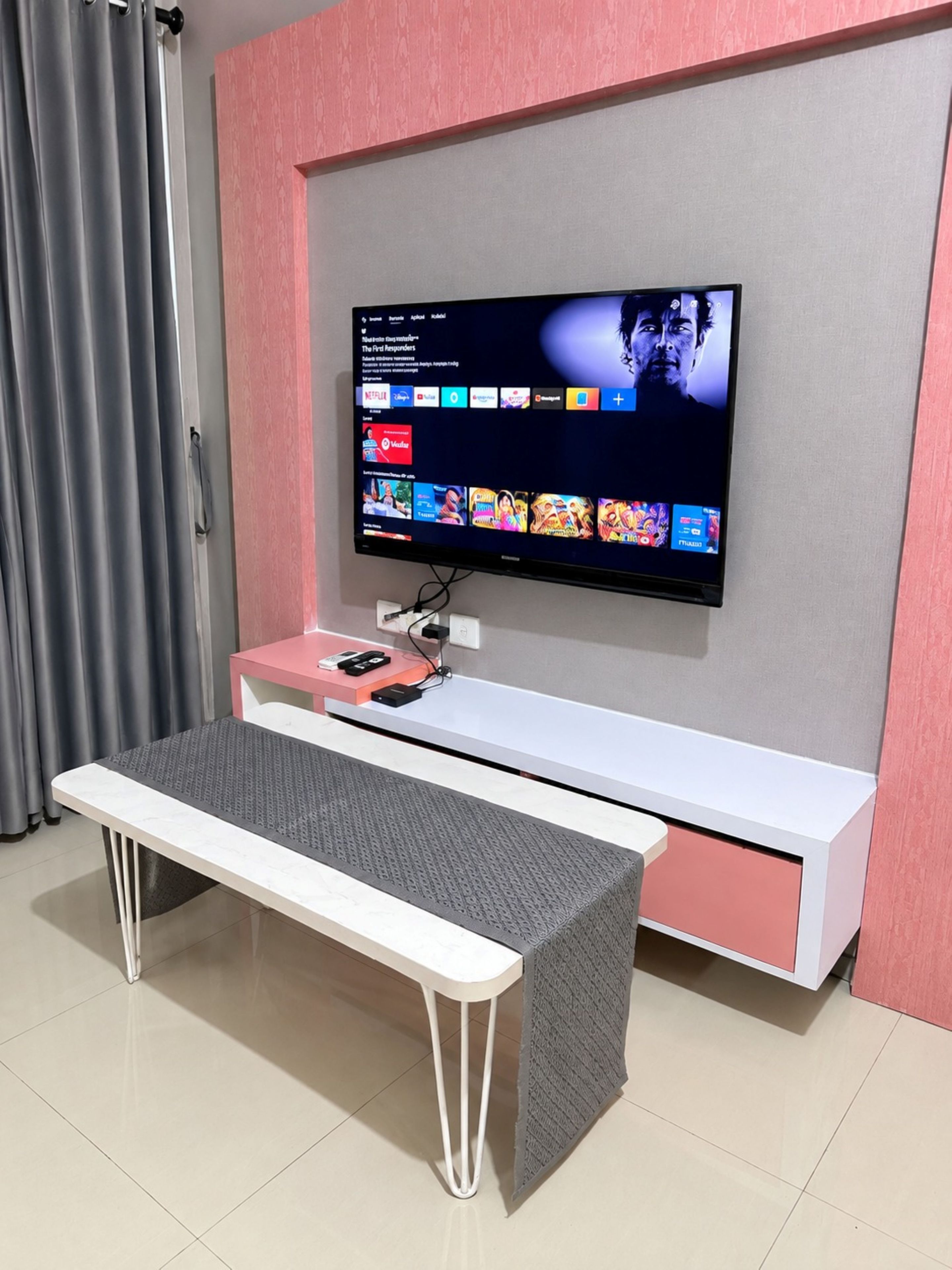A modern living room with a flat-screen TV mounted on a pink wall, a white bench, and a small table with a pink lamp.
