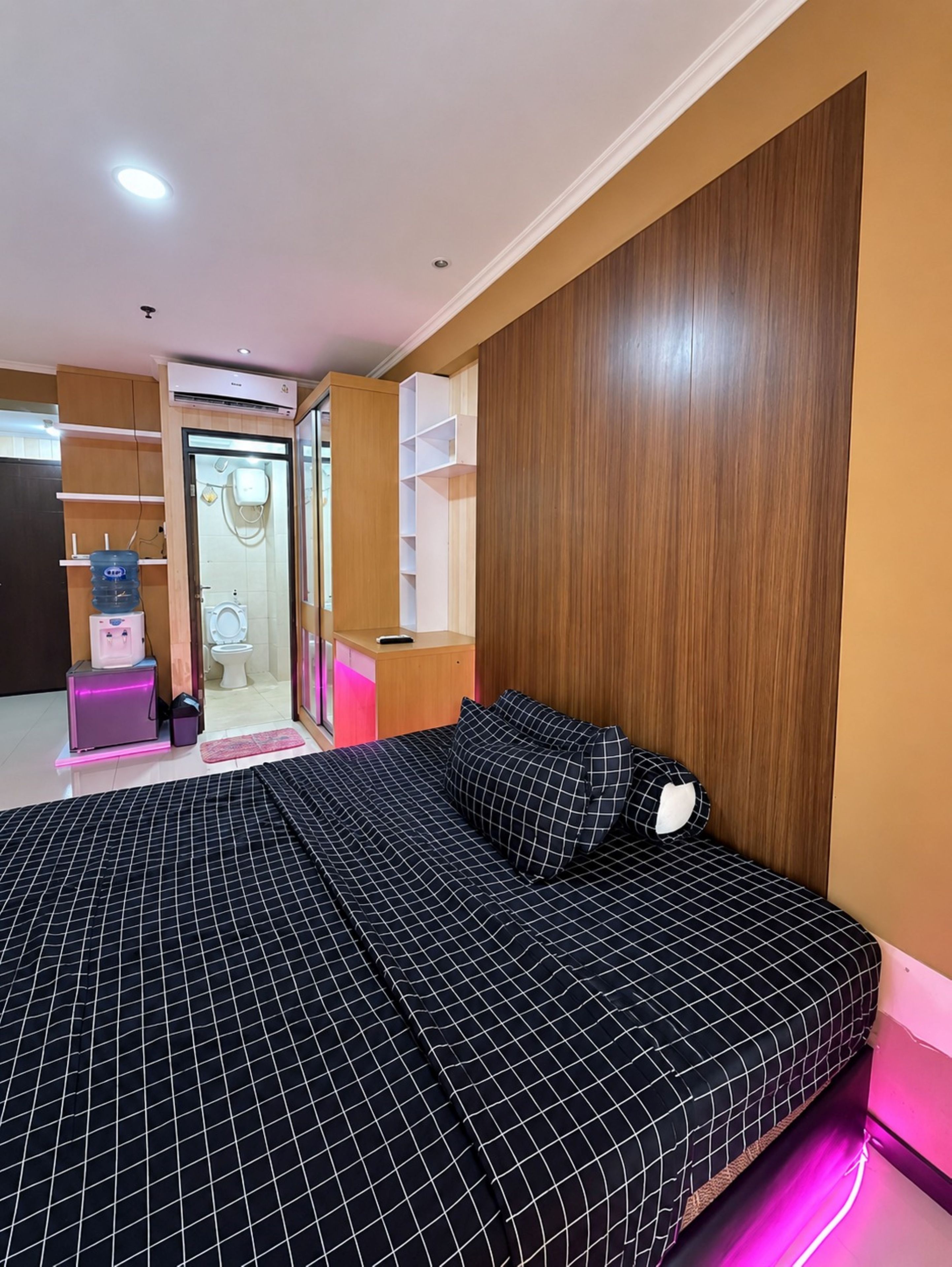 A bedroom with a bed, a wooden headboard, a bathroom with a toilet and sink, and a water dispenser.