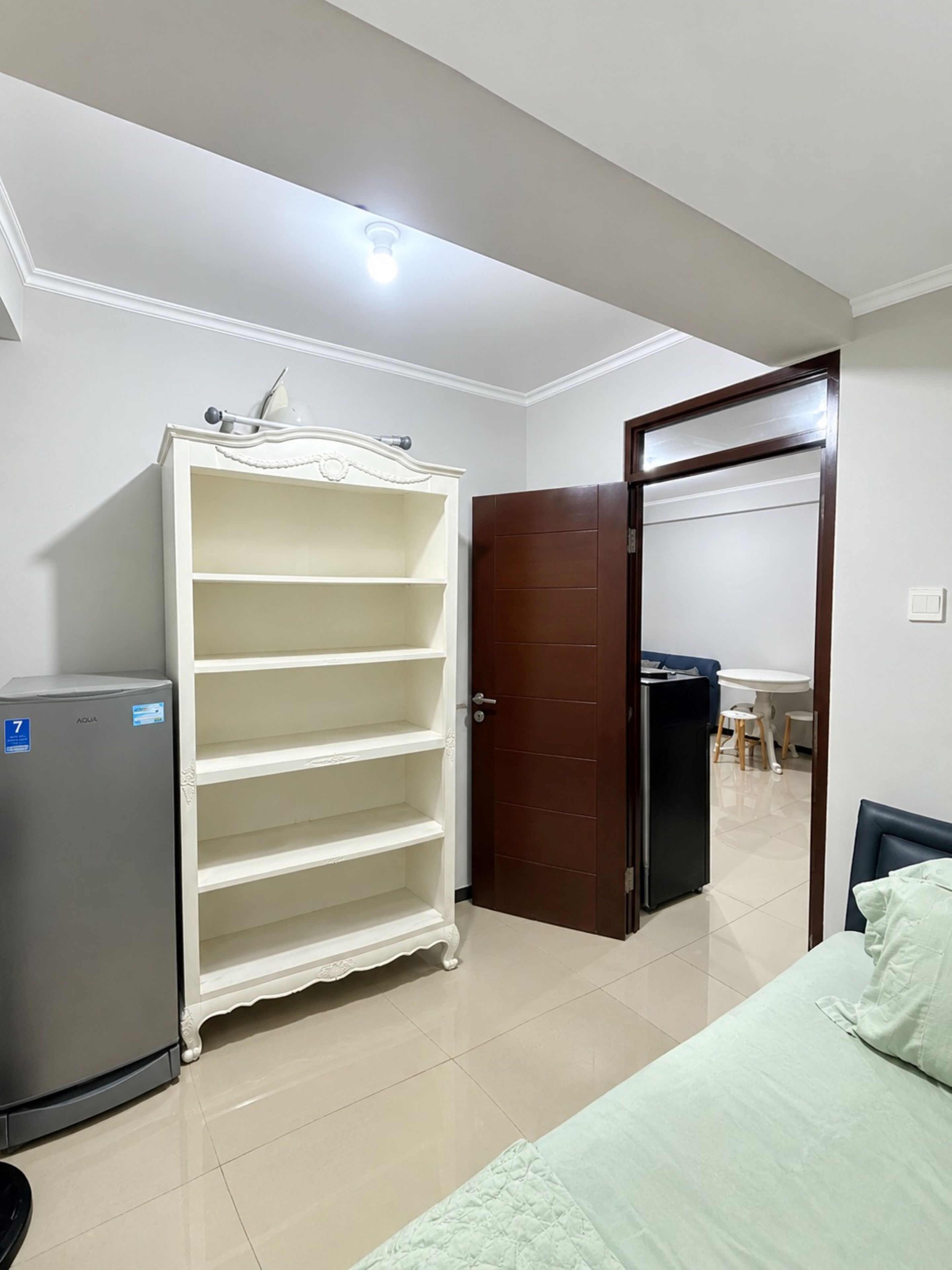 A room with a white wardrobe, a small refrigerator, a bed with a green quilt, and a dining area with a table and chairs.