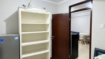A room with a white wardrobe, a small refrigerator, a bed with a green quilt, and a dining area with a table and chairs.