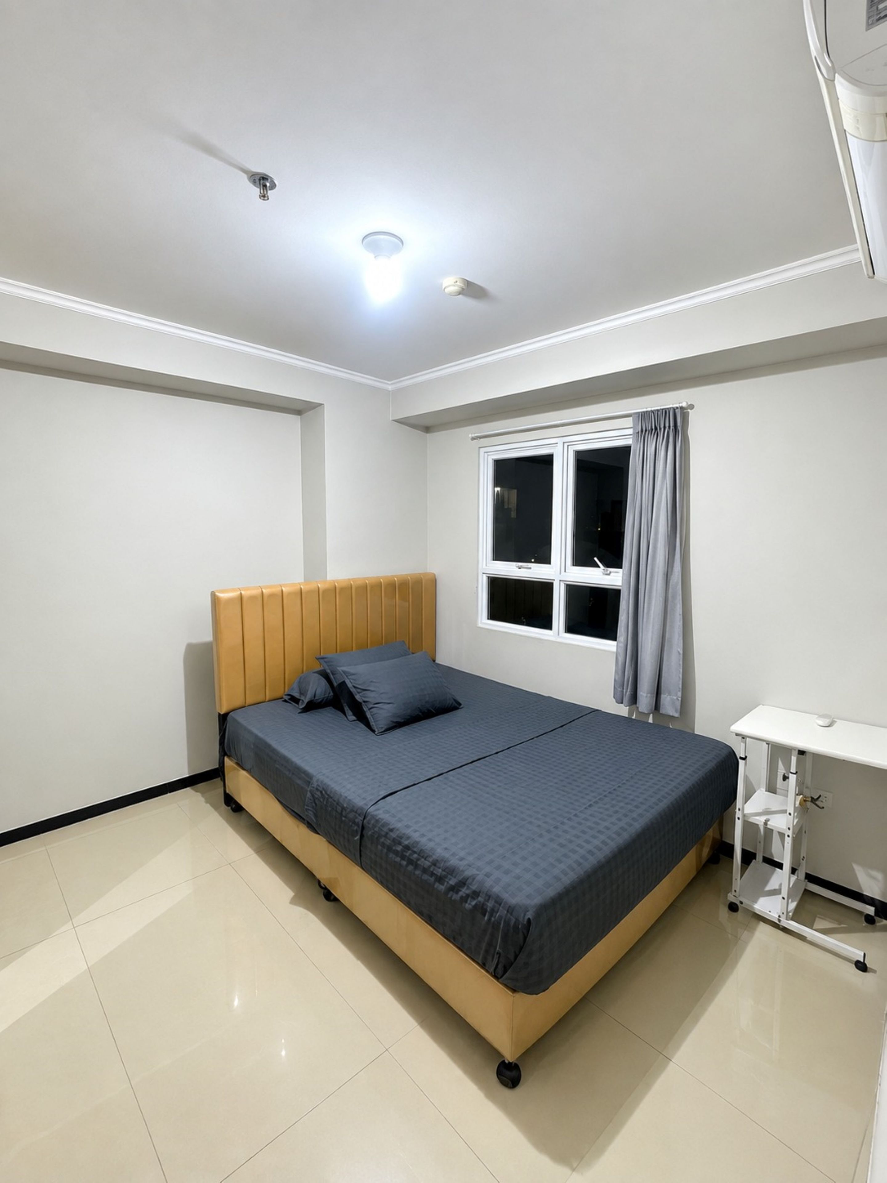 A bedroom with a bed, a window with curtains, a desk, and a ceiling light.