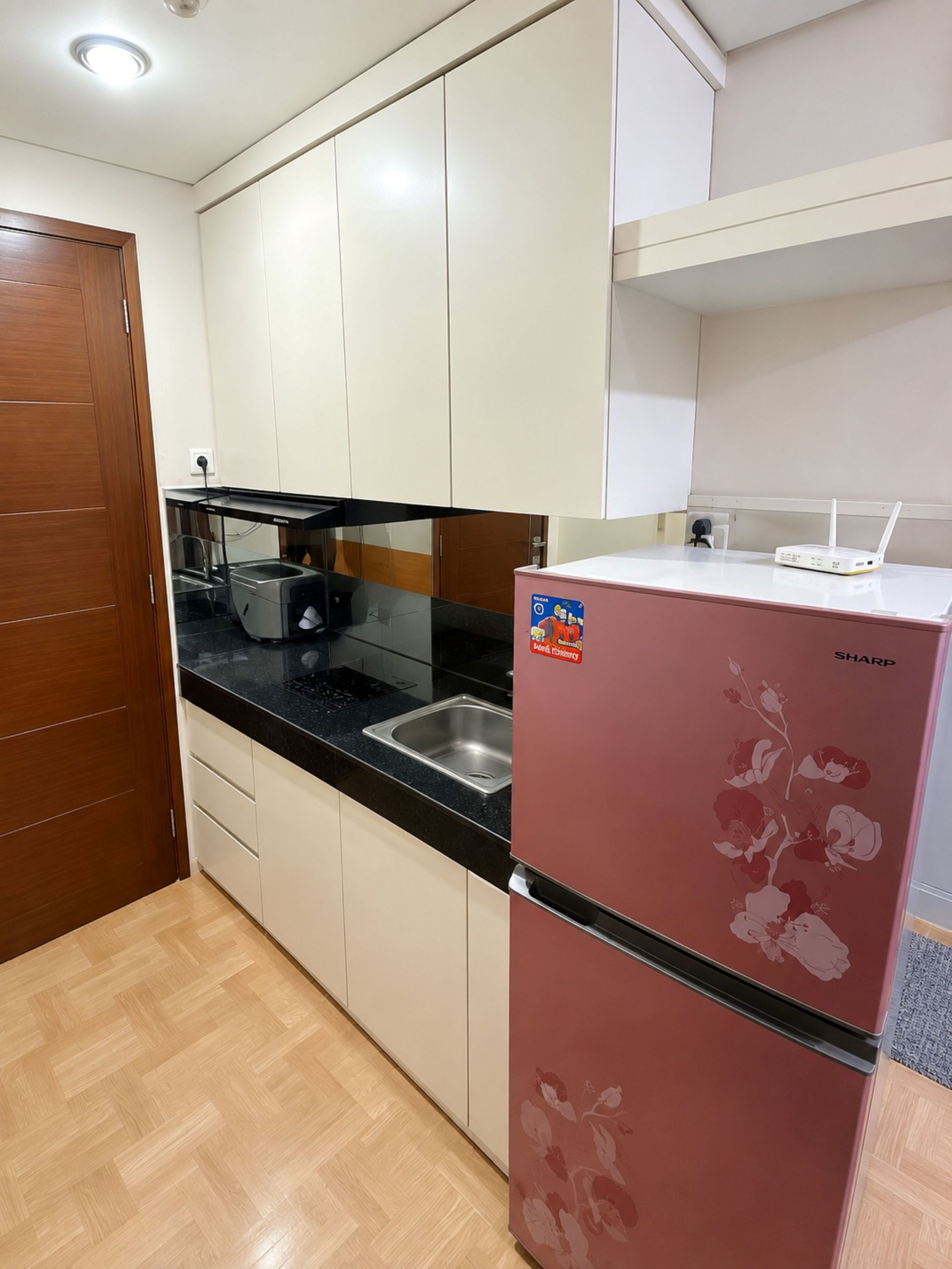 A compact kitchen with white cabinets, a black countertop, a stainless steel sink, a red refrigerator with floral design, and a white appliance on the countertop.