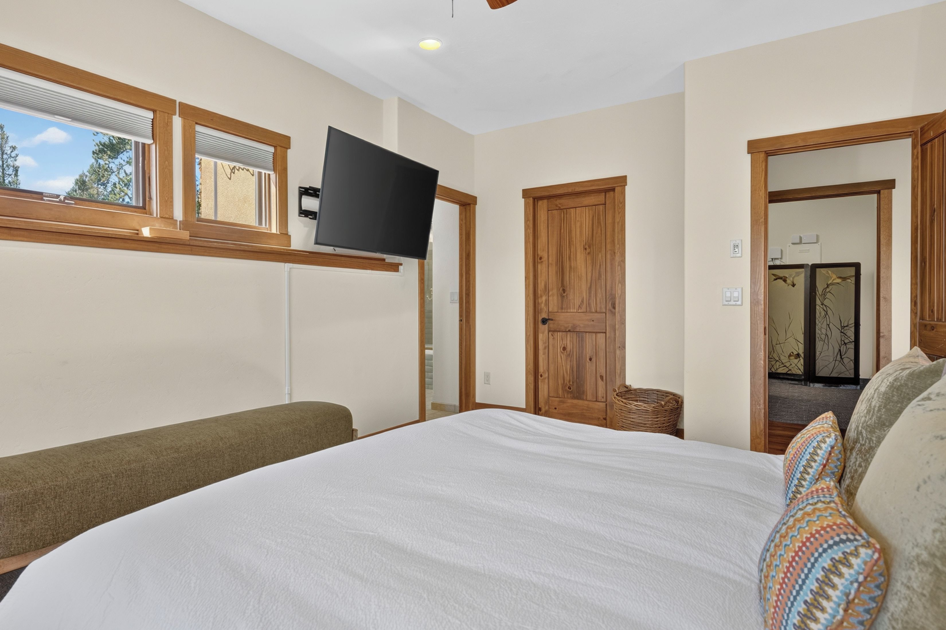 Quarto com cama, televisão na parede, janela com persianas e porta de madeira.