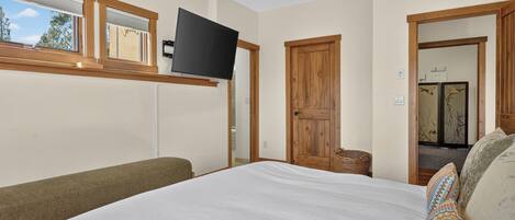 Quarto com cama, televisão na parede, janela com persianas e porta de madeira.