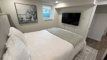 A bedroom with a bed, a flat-screen TV mounted on the wall, a window with blinds, and a patterned rug on the floor.