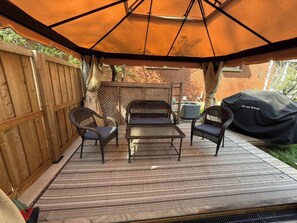 An outdoor patio with a wooden deck, a metal table, two wicker chairs, and a covered area with an umbrella.