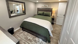 A bedroom with a bed, a bedside table, a lamp, a mirror, and a door. - Modern 2BR Retreat w/ Heated Floors & Gazebo (Brantford)