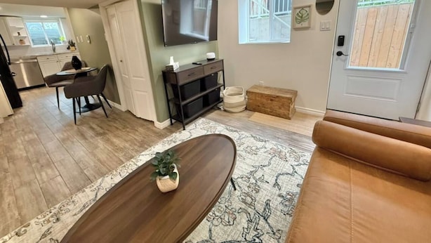 A living room with a sofa, a coffee table, a TV mounted on the wall, and a window with blinds. - Modern 2BR Retreat w/ Heated Floors & Gazebo (Brantford)