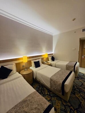 Ein Hotelzimmer mit drei Betten, einem Kopfteil mit integrierter Beleuchtung, einem gemusterten Teppich und einer Tür rechts.