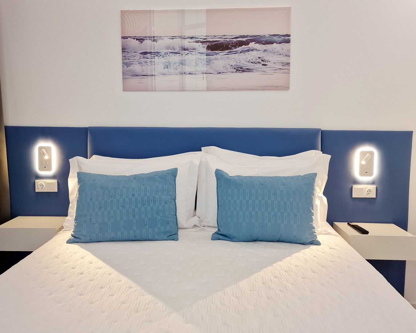 Habitación de hotel con una cama, dos mesitas de noche, una lámpara de pared y una imagen del océano.