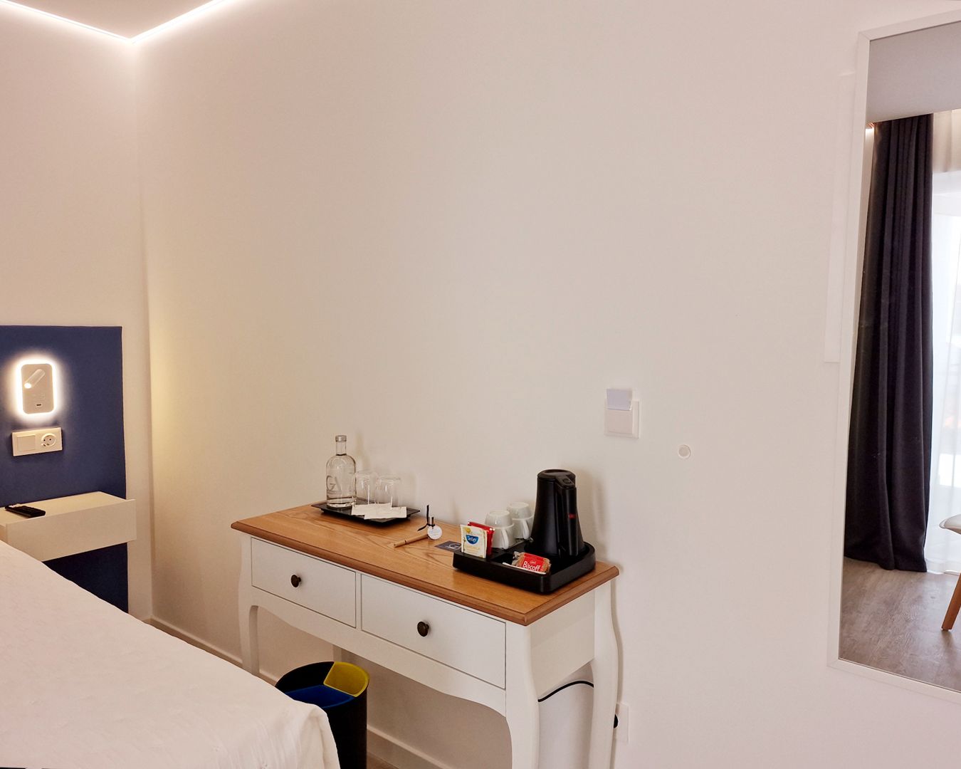 A hotel room with a wooden desk, a bed, a trash bin, and a mirror.