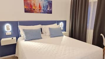 Una cama bien hecha con edredón blanco y almohadas azules, un cuadro de veleros en la pared y una mesita de noche con una lámpara.