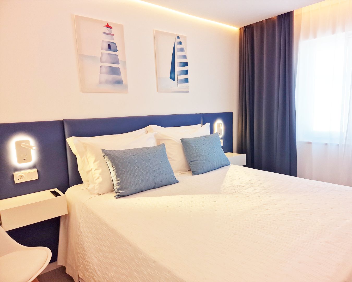 A hotel room with a bed, bedside tables, a nightstand, a chair, and artwork on the wall.