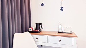 Habitación de hotel con un escritorio de madera, una silla blanca, cafetera, botella de agua y un estante pequeño con objetos decorativos.
