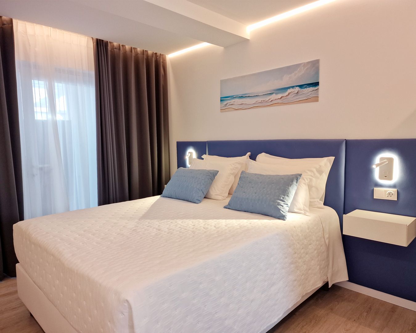 A hotel room with a large bed, a headboard with integrated lighting, a painting of a beach scene, and a bedside table with a lamp.