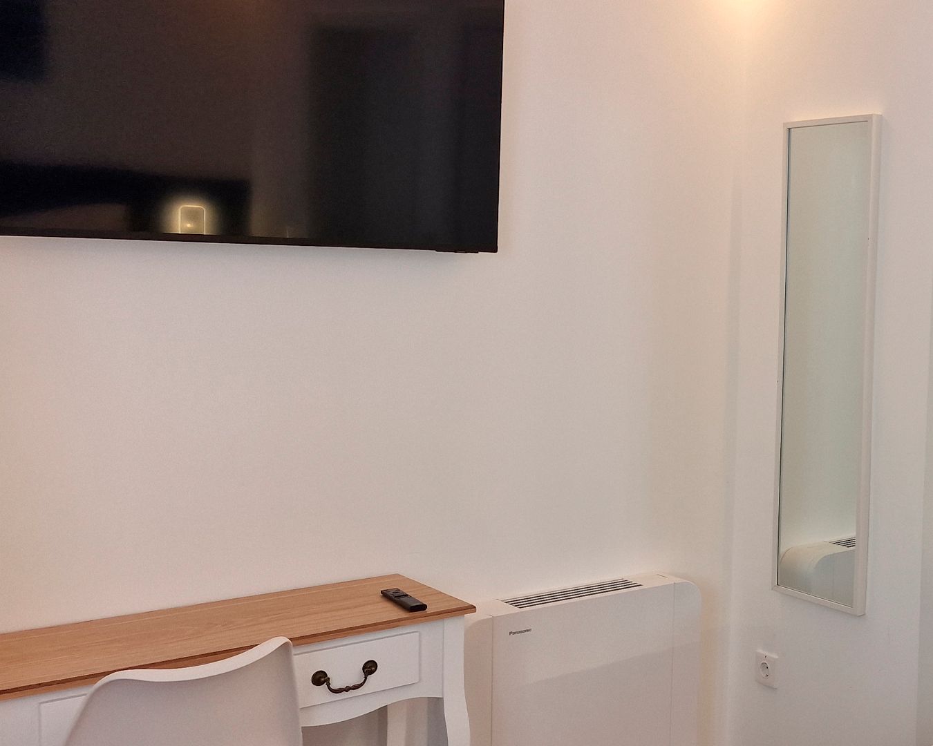 A room with a white desk, a mirror, and a wall-mounted television.